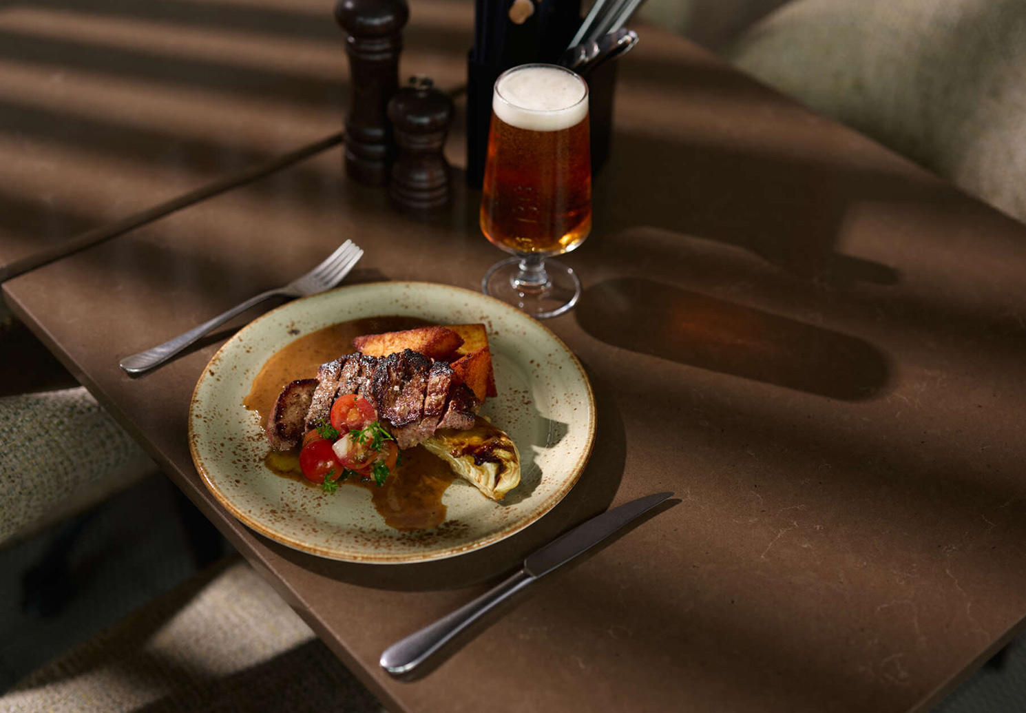 Plate with sliced steak, sauce and side dishes, with a glass of beer on the table.